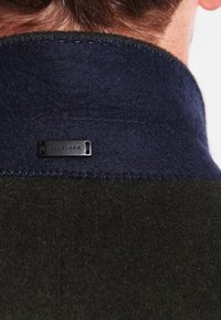 Col en laine mélange vert foncé et bleu marine avec une étiquette noire portant l'inscription "HILFIGER" en relief. Détails de couture visibles le long du bord.