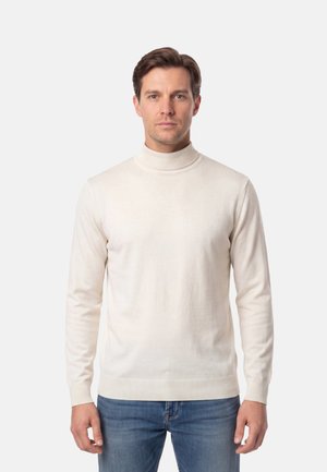 Uomo che indossa un dolcevita color crema e jeans blu, in piedi davanti a uno sfondo chiaro e uniforme.