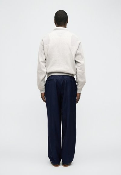 Άνδρας που στέκεται με την πλάτη προς τα εμπρός, φοράει ανοιχτό γκρι πουλόβερ και μπλε σκούρο παντελόνι με λεπτές ρίγες και φαρδιά πόδια, σε απλό φόντο.