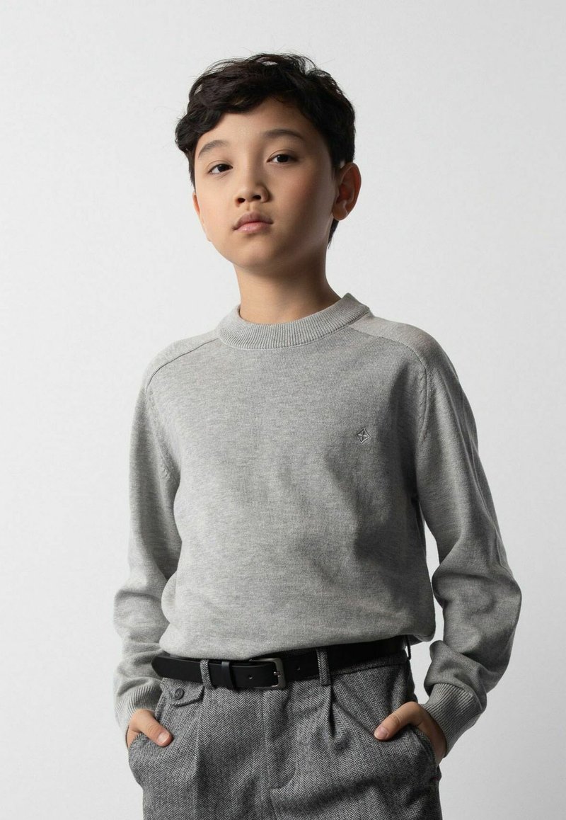 Szary dzianinowy sweter z okrągłym dekoltem i prążkowanymi mankietami, zestawiony z szarymi spodniami w jodełkę. Na piersi znajduje si�ę małe logo.