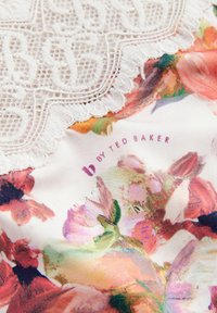 Textília s kvetinovým vzorom, ktorá obsahuje viacfarebné odtiene s bielou čipkovou lemovkou na vrchu a logom "B by Ted Baker" v purpurovej farbe.