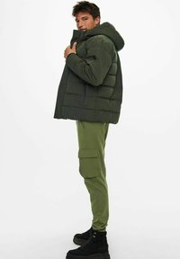 Dark green puffer jacket with a hood, featuring a quilted design and zip closure. Paired with olive green cargo pants and black boots.