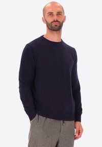 Uomo con barba che indossa un maglione blu scuro e pantaloni grigi, in piedi con una mano in tasca su uno sfondo uniforme.