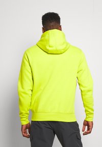 Man som vänder sig bort klädd i en ljusgul hoodie och mörkgrå cargobyxor mot en enfärgad ljus bakgrund.