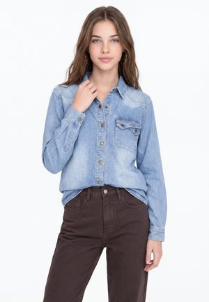 Jeune femme aux longs cheveux bruns portant une chemise en jean bleu clair et un pantalon marron foncé, debout devant un fond blanc.