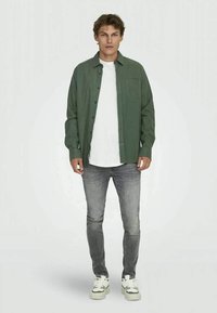 Camisa verde de manga longa, usada por cima de uma t-shirt branca, combinada com jeans cinzentos e sapatilhas brancas. Apresenta um bolso no peito e um corte descontraído.