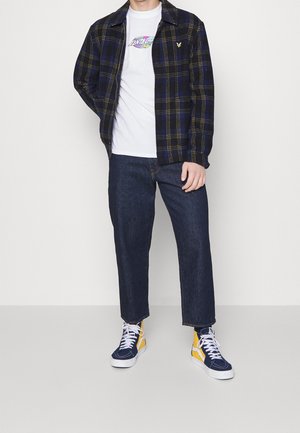 Camisa de franela a cuadros en navy y negro sobre una camiseta gráfica blanca, combinada con unos jeans rectos de mezclilla oscura y zapatillas con acentos amarillos.