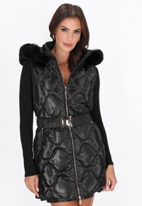 Vest negru matlasat cu glugă, având margine din blană sintetică, fermoar frontal și talie cu brâu. Suprafață netedă cu modele în diamant.