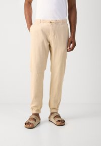 Beige linnen broek met een elastische tailleband en trekkoord, met zijzakken en taps toelopende boorden, gecombineerd met beige sandalen.