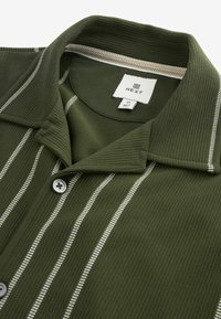 Chemise en maille côtelée vert olive avec rayures blanches verticales, devant boutonné, col à revers, et étiquette de marque "NEXT" visible à l'intérieur du col.