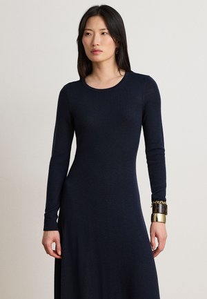Femme aux cheveux noirs et raides portant une robe ajustée à manches longues bleu marine foncé et des bracelets volumineux à son poignet gauche sur un fond uni.