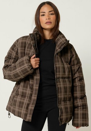 Femme portant une doudoune marron à carreaux par-dessus une chemise noire, tenant le col de la veste d'une main devant un fond clair uni.