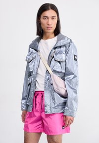 Silver waterproof jacket with multiple front pockets, grey hood, and a small black logo, paired with bright pink shorts and a light pink bag.