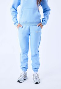 Sudadera azul claro con un bolsillo frontal y pantalones de chándal a juego que presentan un logo bordado. Puños redondeados y un corte relajado. Zapatillas plateadas.