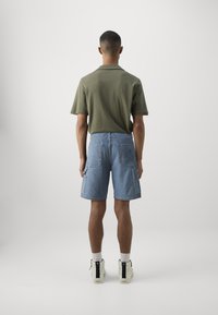 Polo shirt vert olive à manches courtes associé à un short cargo en denim bleu clair avec deux poches plaquées et un détail de logo. Baskets blanches avec accents noirs.