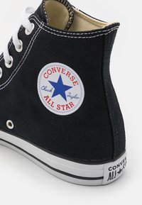 Černé plátěné vysoké tenisky s bílými tkaničkami, šitím a gumovou podrážkou. Obsahuje kruhové logo s modrou hvězdou a textem "Converse All Star."