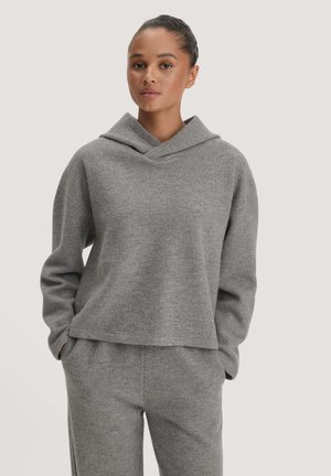 Jonge vrouw draagt een grijze hoodie en bijpassende joggingbroek met handen in de zakken tegen een effen lichte achtergrond.
