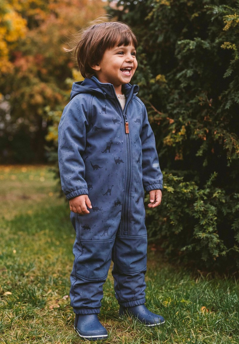 Enfant souriant en combinaison de pluie bleue à motifs de dinosaures et bottes se tient sur l'herbe près d'arbres à feuilles persistantes par temps nuageux.