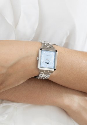 Silberne Metalluhr mit blauem rechteckigem Zifferblatt, silbernen Stundenmarkierungen und einem Gliederarmband, getragen an einem hellen Hautton.