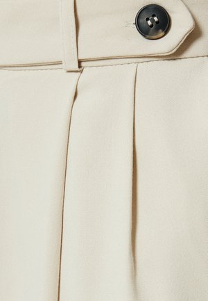 Kerman värinen taitteellinen kangas, jossa on teksturoitu pinta, varustettuna mustalla napilla ja tikattuilla yksityiskohdilla vyötärön ympärillä.