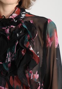Blusa negra transparente con patrones florales coloridos en rosa y verde azulado. El escote con volantes y los detalles de tela en capas añaden textura.