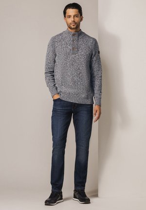 Warmer blauer Strickpullover mit hohem Kragen und drei Holzknöpfen. Kombiniert mit dunkelblauen Jeans und schwarzen Turnschuhen.