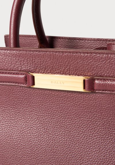 Burgundia nahast käekott, millel on tekstureeritud viimistlus, ja kuldse tooni metallist detail, mis kannab brändi nime "BALLY" horisontaalsel plaadil.