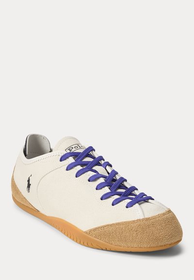 Baskets basses blanches et beige avec des lacets bleus et un logo Polo noir sur le côté, montrées légèrement en angle sur fond blanc.