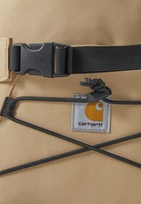Tecido bege com uma superfície texturizada, apresentando uma fivela preta, cordões elásticos e uma etiqueta cinza com um logotipo amarelo.