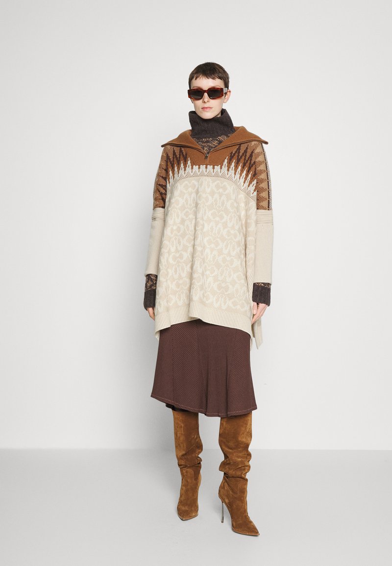 Übergroßer beigefarbener und brauner gestrickter Poncho mit geometrischen Mustern, kombiniert mit einem braunen, plissierten Rock, ergänzt durch kniehohe, braune Wildlederstiefel.