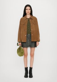 Giacca in suede marrone, top in maglia verde, gonna in pelle nera con zip e stivaletti neri alla caviglia. Tiene una borsa verde.