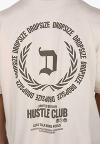 Fehér pamut póló, amelynek hátulján egy nagy grafikával díszített nyomat található. A szöveg tartalmazza a "HUSTLE CLUB" feliratot és egy díszes babérkoszorút sötétszürke színben.