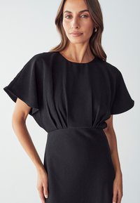 Robe noire avec de larges manches fluides et une taille ajustée. Fabriquée dans un tissu lisse, avec de subtils détails de plis au niveau du buste.