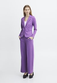 Paars op maat gemaakte blazer met bijpassende brede broek, gemaakt van soepele stof. Beschikt over voorzakken en gestructureerde revers. Zwarte hakken maken de look compleet.