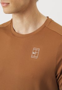 T-shirt atlético castanho feito de tecido texturizado, com o logótipo branco da Nike dentro de um contorno retangular na área do peito esquerdo.