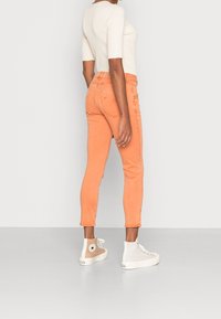 Jeans slim fit taille haute, de couleur pêche avec une texture lisse, dotés de poches arrière et associés à des sneakers montants blanches et beiges.