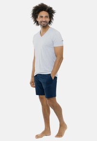 Graues T-Shirt mit V-Ausschnitt und marineblaue Shorts mit Seitentaschen. Das Modell steht barfuß, lässig, mit lockigem Haar und Bart.