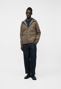 Άνδρας που στέκεται φορώντας καφέ μπουφάν με κουκούλα, μπλε πουκάμισο, σκούρα τζιν φαρδιάς γραμμής με κόκκινες ραφές και μαύρα δερμάτινα παπούτσια.