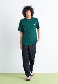 Dark green short-sleeve t-shirt with a round neckline, paired with black athletic pants and black shoes with pink accents. Smooth fabric texture.