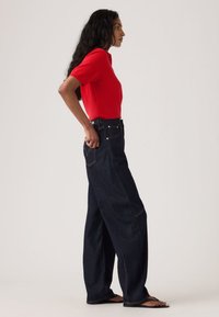 Κόκκινο τοπ με κοντά μανίκια συνδυασμένο με ψηλόμεσα σκούρα τζιν παντελόνια φαρδιάς γραμμής, με πλαϊνές τσέπες και λεπτομέρειες με αντίθετη ραφή.