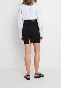 Falda de mezclilla negra con una gran abertura en la parte posterior, con cintura de altura media y bolsillos. Combinada con una camisa de botones azul clara y sandalias negras.