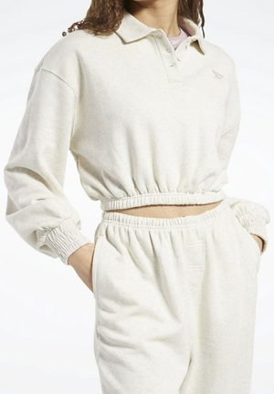 Vrouw draagt een crèmegekleurde cropped sweatshirt met elastische zoom en bijpassende high-waist joggingbroek met handen in de zakken.