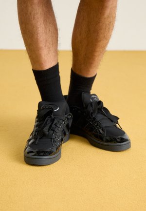 Jambes portant des chaussettes noires courtes et des baskets noires brillantes, debout sur un sol recouvert d’un tapis jaune, sur un fond uni.
