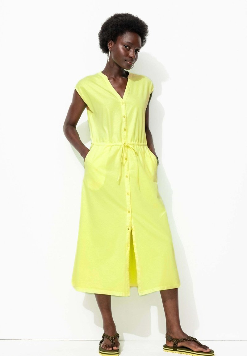 Robe jaune à boutons avec un décolleté en V, manches courtes, cordon de serrage à la taille et poches latérales, en tissu léger. Sandales à lanières.