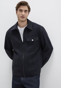 Dunkelblaues Zip-Up-Jacket aus strukturiertem Stoff, mit einer einzelnen Brusttasche und großem Kragen. Über einem weißen T-Shirt getragen.