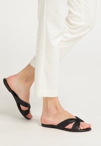 Sandali neri in pelle con cinturini attorcigliati, suola piatta e design aperto. Indossati con pantaloni bianchi, mostrano uno stile minimalista.