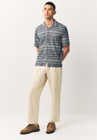 Kurzärmeliges Hemd mit Knöpfen in Marineblau mit weißen geometrischen Mustern, kombiniert mit cremefarbenen Hosen und braunen Wildlederschuhen.