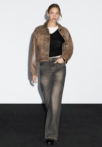 Braune Leder-Cropped-Jacke über einem schwarzen V-Ausschnitt-Shirt, kombiniert mit grauen, weit geschnittenen Jeans und schwarzen Plateau-Schuhen. Schlichtes Design und Texturen.