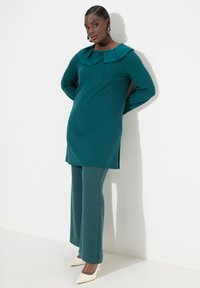 Tunika turchese con un ampio colletto pieghettato, abbinata a pantaloni a gamba larga coordinati e scarpe bianche con punta. Tessuto morbido; silhouette aderente.