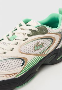 Chaussure avec tige en mesh blanc agrémentée d'accents verts et dorés, languette rembourrée, lacets ronds et semelle en caoutchouc noire. Logo Lacoste sur le côté.